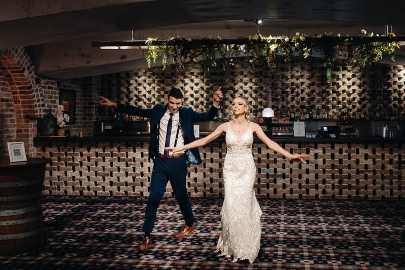 Courtney the bride and Cameron the groom dance together on the reception stage at Sandstone Point Hotel — Cellar, with a brick wall and bar in the background.