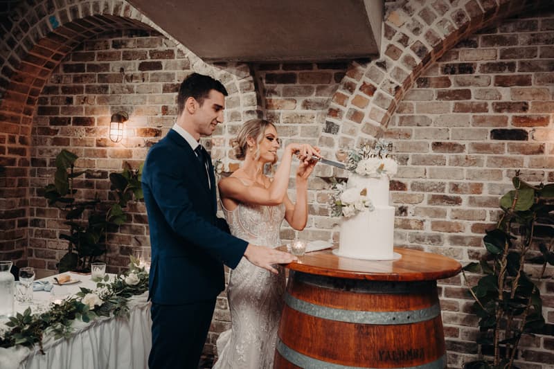Courtney the bride and Cameron the groom cut their wedding cake on a wooden barrel at Sandstone Point Hotel — Cellar, with a brick wall and plants in the background.