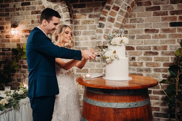Courtney the bride and Cameron the groom cut their wedding cake on a wooden barrel table at Sandstone Point Hotel — Cellar, with a brick wall backdrop and decorative plants.