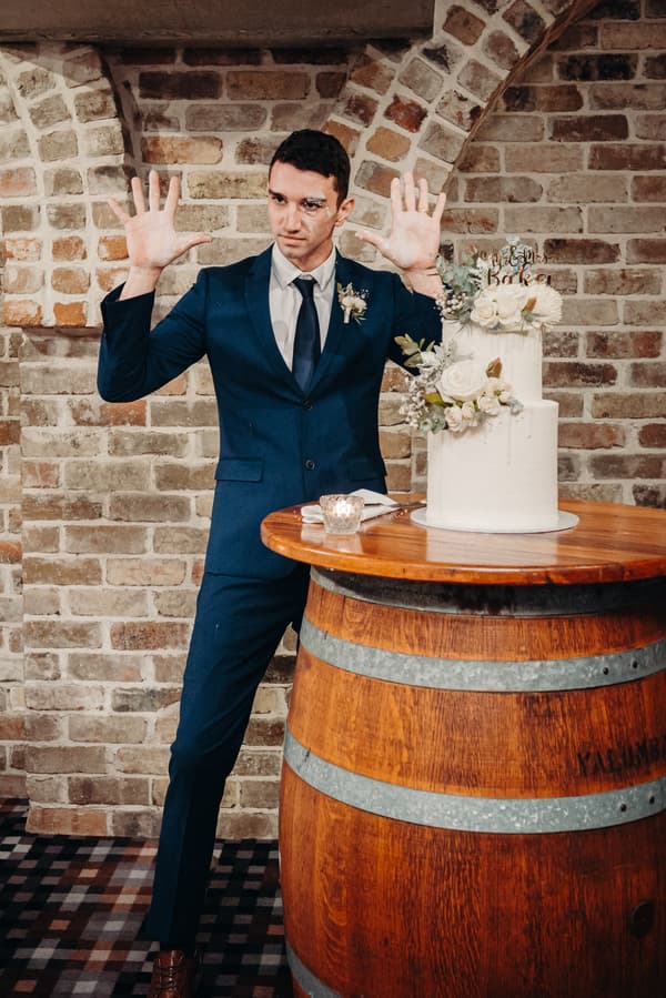 The groom Cameron stands next to a two-tier white wedding cake on a wooden barrel table at Sandstone Point Hotel — Cellar, with his hands raised and some cake on his face.