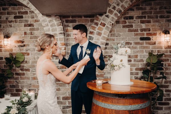 Courtney the bride and Cameron the groom stand by a wooden barrel table with a two-tier white wedding cake decorated with flowers at Sandstone Point Hotel — Cellar.