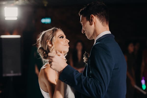 Courtney and Cameron share a dance on the reception stage at Sandstone Point Hotel — Cellar, with guests blurred in the background.