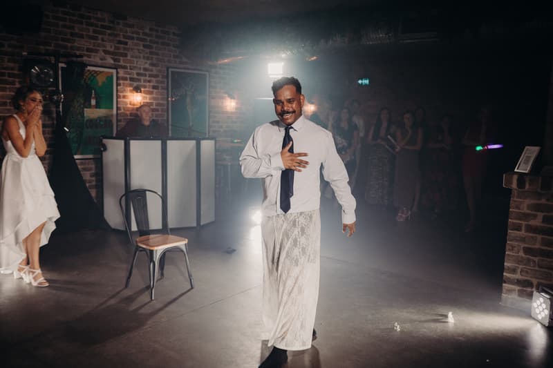 The groom Cameron dances wearing a white shirt, black tie, and a white lace skirt at the Sandstone Point Hotel — Cellar reception stage, while the bride Courtney watches with her hands covering her mouth in surprise or amusement. Guests stand in the background near a brick wall and DJ booth.