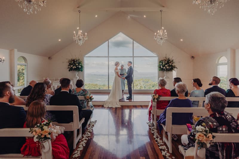 Courtney and Liam stand at the altar holding hands during their wedding ceremony at Tiffany's Maleny — Chapel, with an officiant between them and guests seated on both sides of the aisle.