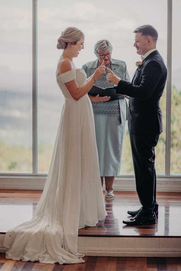 Courtney and Liam exchange rings during their wedding ceremony at Tiffany's Maleny — Chapel, with the officiant standing behind them.