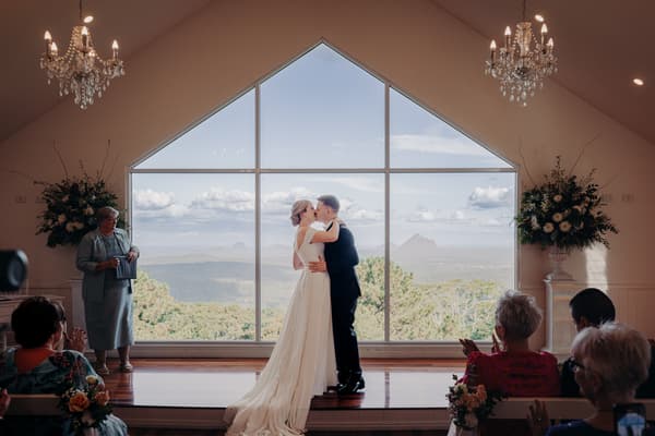 Bride Courtney and groom Liam share a kiss at the altar during their wedding ceremony at Tiffany's Maleny — Chapel, with guests seated and applauding in the foreground and a large window showing a scenic mountain view behind them.