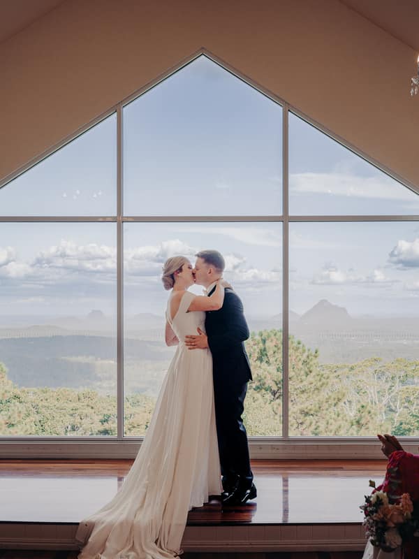 Bride Courtney and groom Liam share a kiss at the altar during their wedding ceremony at Tiffany's Maleny — Chapel, with guests seated and applauding in the foreground and a large window showing a scenic mountain view behind them.