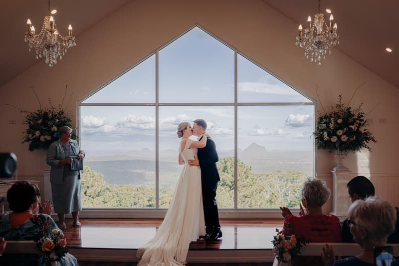 Bride Courtney and groom Liam share a kiss at the altar during their wedding ceremony at Tiffany's Maleny — Chapel, with guests seated and applauding in the foreground and a large window showing a scenic mountain view behind them.