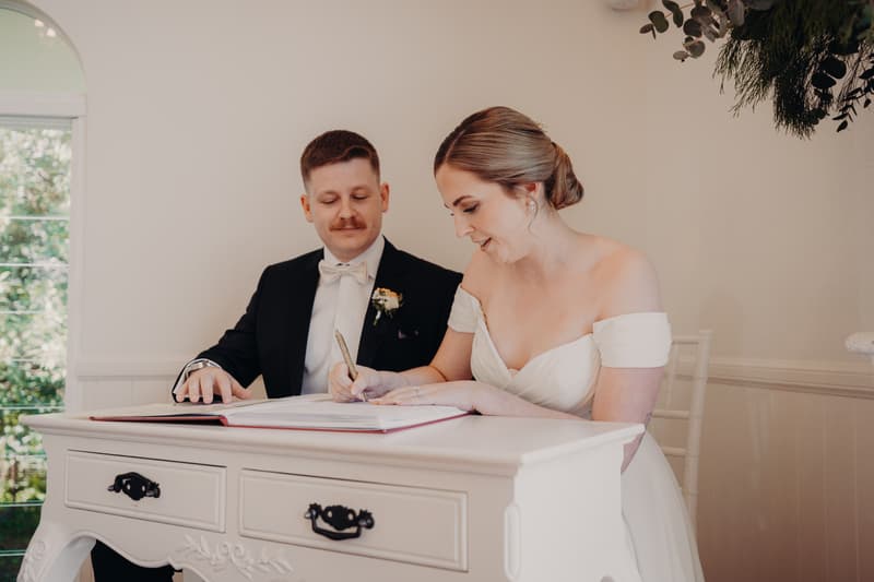 Courtney the bride signs the wedding register at Tiffany's Maleny — Chapel while Liam the groom looks on.