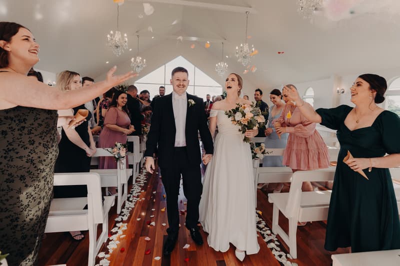 Courtney the bride and Liam the groom walk down the aisle at Tiffany's Maleny — Chapel as guests throw flower petals.