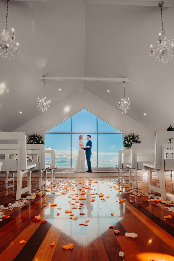 The bride Courtney and groom Liam stand facing each other holding hands inside Tiffany's Maleny chapel with white benches, chandeliers, and scattered orange and white flower petals on the wooden floor.