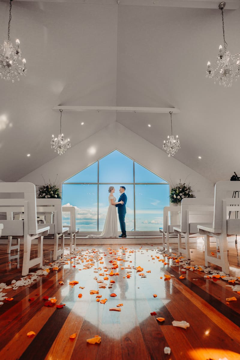The bride Courtney and groom Liam stand facing each other holding hands inside, chandeliers, and scattered orange and white flower petals on the wooden floor. ABIA 2018 finalist wedding photography by She Said Yes.