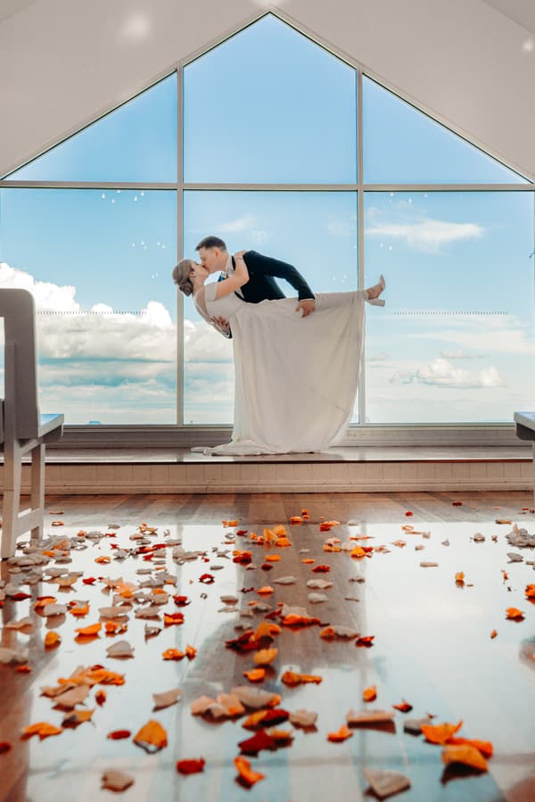The bride Courtney and groom Liam pose in a dip kiss in front of a large triangular window at Tiffany's Maleny, with scattered orange and white flower petals on the wooden floor.