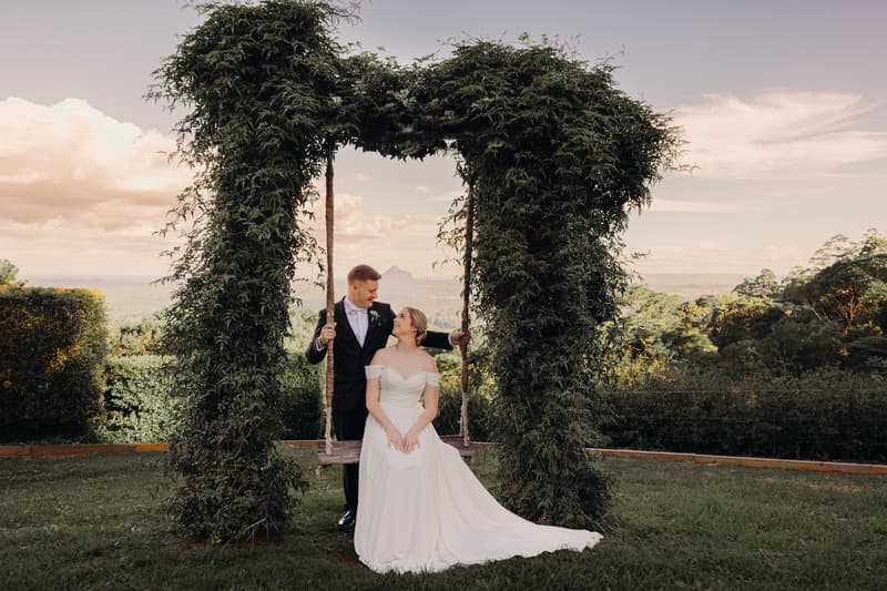 Courtney and Liam pose together on a wooden swing framed by a leafy arch at Tiffany's Maleny, with a scenic landscape in the background.
