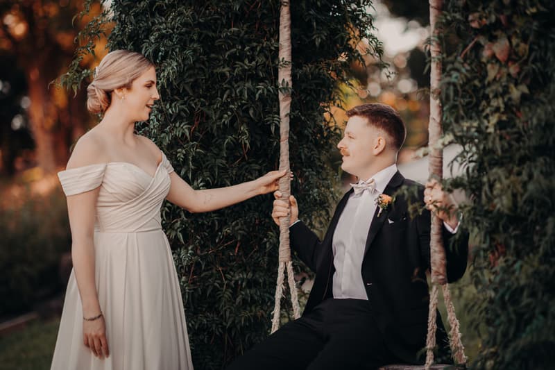 Courtney the bride stands beside Liam the groom who is seated on a rope swing at Tiffany's Maleny, surrounded by greenery.