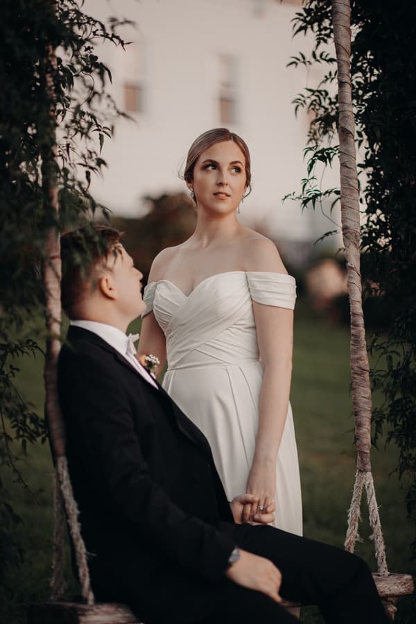 Courtney the bride in an off-shoulder white wedding dress stands holding hands with Liam the groom, who is seated on a swing wrapped with fabric, at Tiffany's Maleny.
