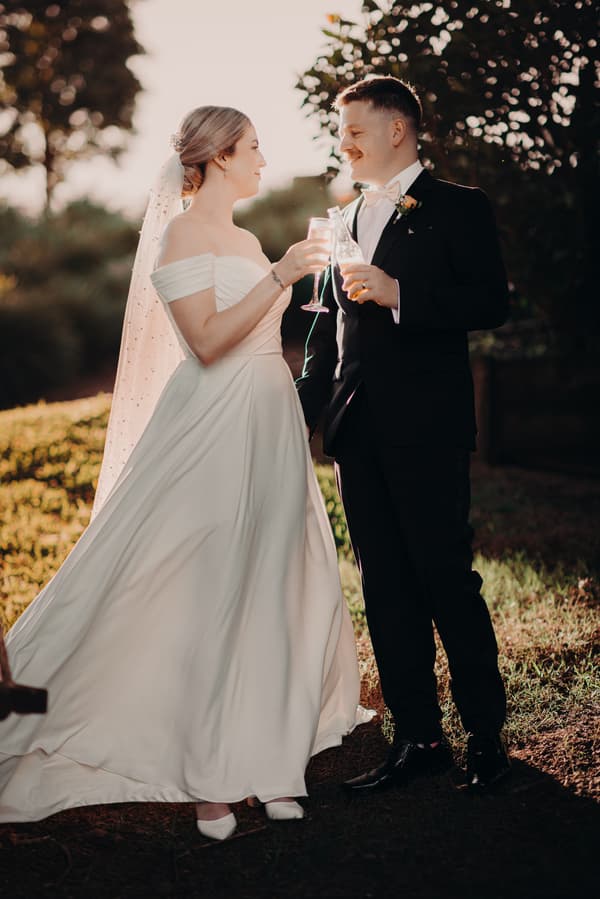 Courtney the bride and Liam the groom stand outdoors at Tiffany's Maleny, holding drinks and facing each other during their couple portraits session.
