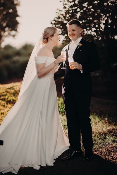Courtney the bride and Liam the groom stand outdoors, holding drinks and smiling at each other during their couple portraits session. Engagment party by She Said Yes.