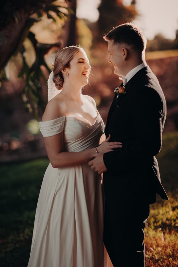 Courtney the bride and Liam the groom stand facing each other outdoors at Tiffany's Maleny during their couple portraits session, with Courtney wearing an off-shoulder wedding gown and Liam in a dark suit with a boutonniere.