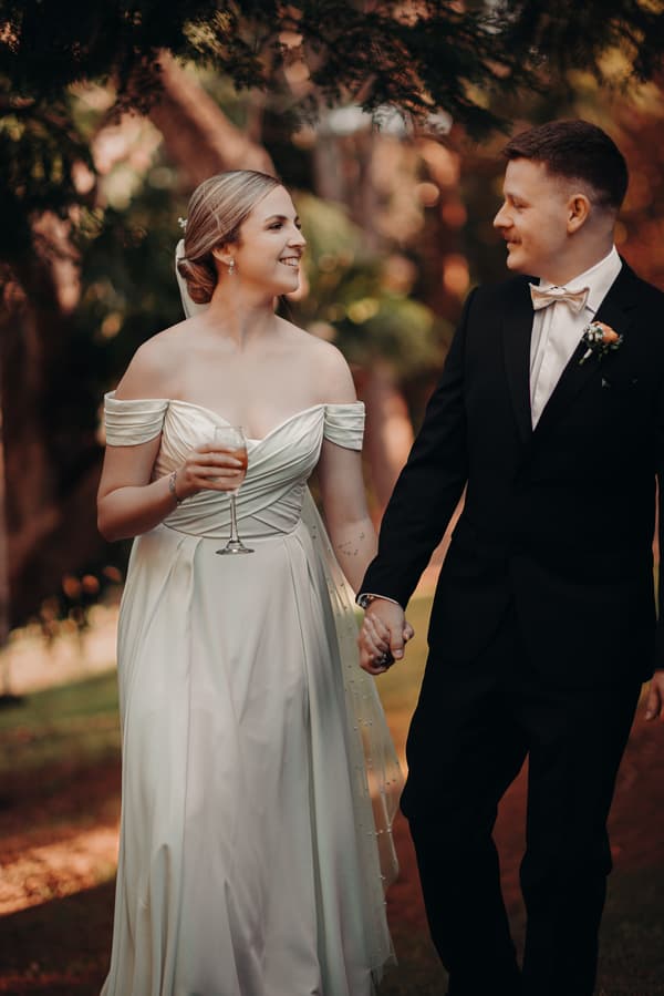 Courtney, the bride, in an off-shoulder white wedding gown holding a glass, and Liam, the groom, in a black suit with a bow tie and boutonniere, walk hand in hand outdoors at Tiffany's Maleny.