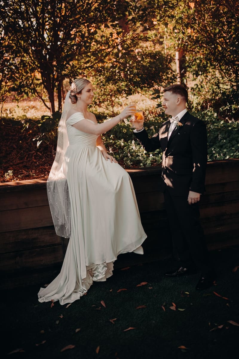 Courtney the bride in a white wedding gown and veil sits on a wooden ledge while Liam the groom in a black suit and bow tie stands facing her, both holding glasses of amber-colored drink in an outdoor garden setting. Sunset wedding portraits Brisbane by She Said Yes.