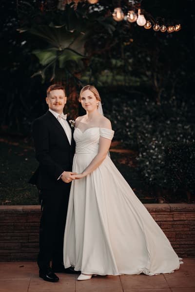 Courtney the bride and Liam the groom pose together holding hands outdoors at Tiffany's Maleny during their couple portraits session. Courtney wears an off-shoulder white wedding gown and veil, while Liam wears a black suit with a white bow tie and boutonniere. String lights hang above them and greenery is visible in the background.