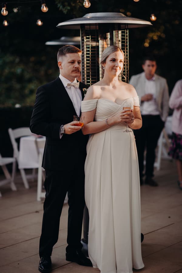 The bride Courtney in an off-shoulder white wedding gown and the groom Liam in a black suit with a bow tie stand together holding drinks near a patio heater at Tiffany's Maleny — Quartyard during the reception.