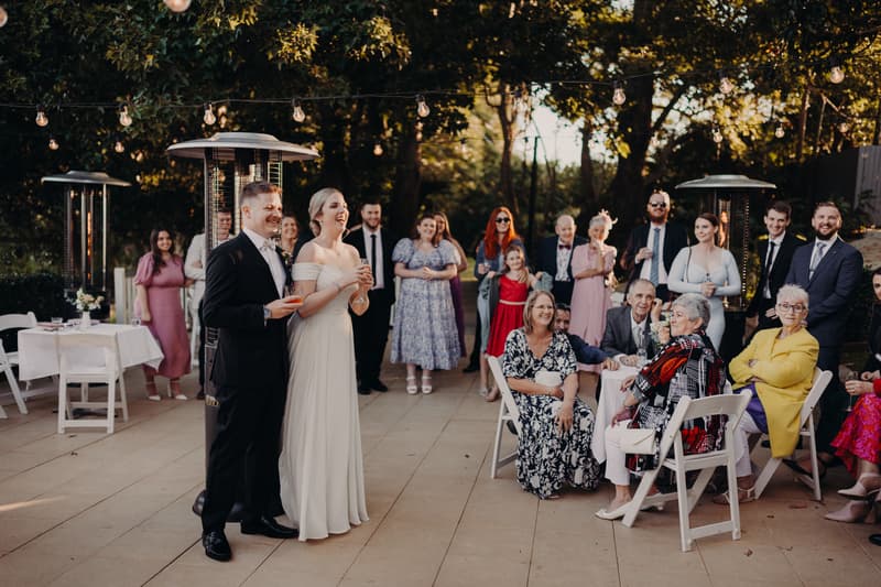 Bride Courtney and groom Liam stand together holding drinks on the reception stage at Tiffany's Maleny — Quartyard, surrounded by seated and standing guests watching them.