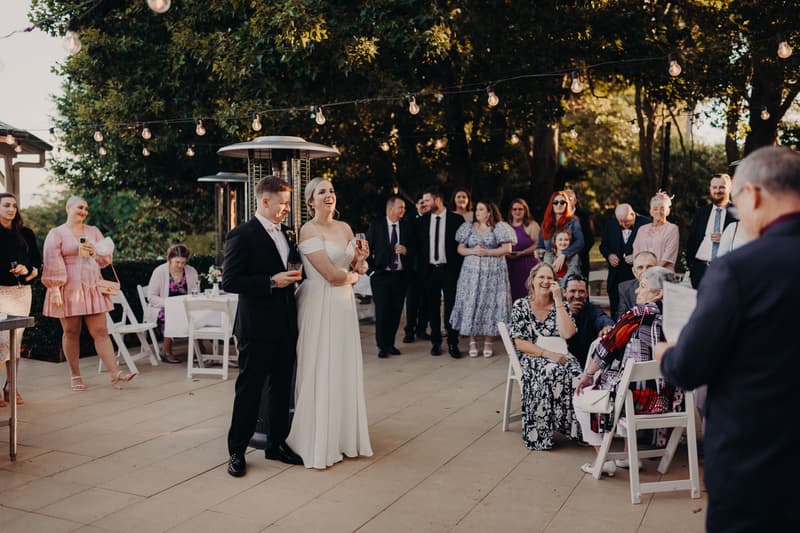 Bride Courtney and groom Liam stand together holding drinks while guests listen to a speech at Tiffany's Maleny — Quartyard reception.