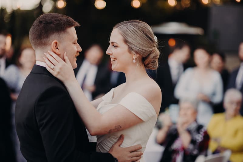 Courtney the bride and Liam the groom share a close moment dancing at the reception stage at Tiffany's Maleny — Quartyard, with guests blurred in the background.