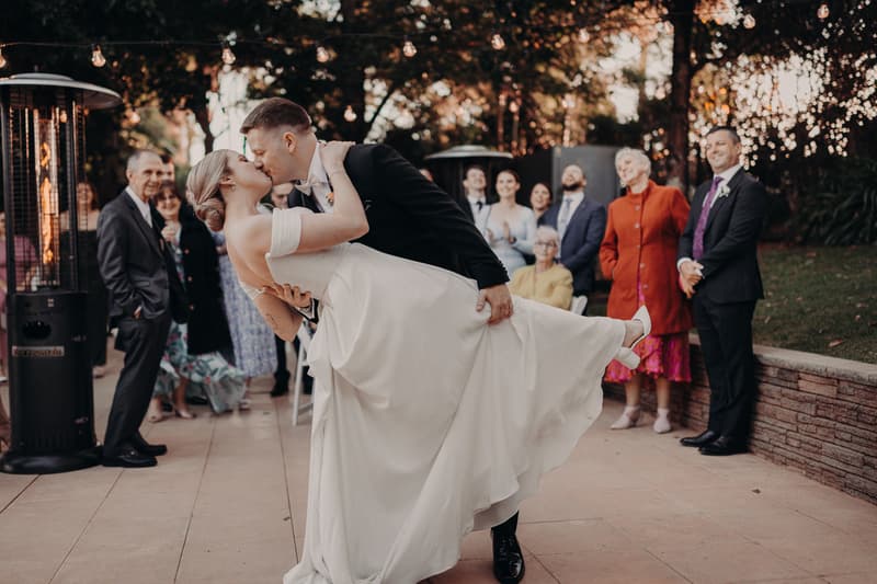 The groom Liam dips the bride Courtney for a kiss on the reception stage at Tiffany's Maleny — Quartyard, while guests watch and smile in the background.