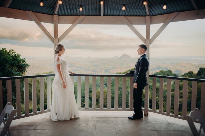 Courtney the bride and Liam the groom stand facing each other on a covered balcony at Tiffany's Maleny with a scenic view of hills and trees in the background.