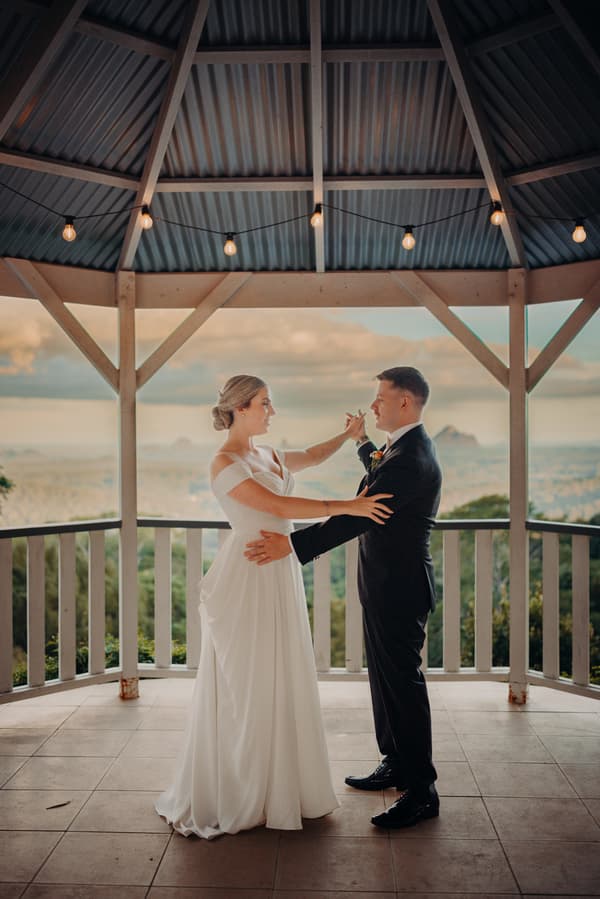 Courtney and Liam pose together under a gazebo at Tiffany's Maleny, with Courtney in a white wedding gown and Liam in a black suit, holding hands and facing each other.