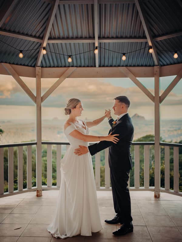 Courtney and Liam pose together under a gazebo at Tiffany's Maleny, with Courtney in a white wedding gown and Liam in a black suit, holding hands and facing each other.