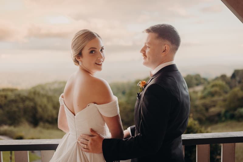 Courtney the bride and Liam the groom pose together on a balcony at Tiffany's Maleny, with Courtney looking back towards the camera and Liam facing her, dressed in a tuxedo with a boutonniere.