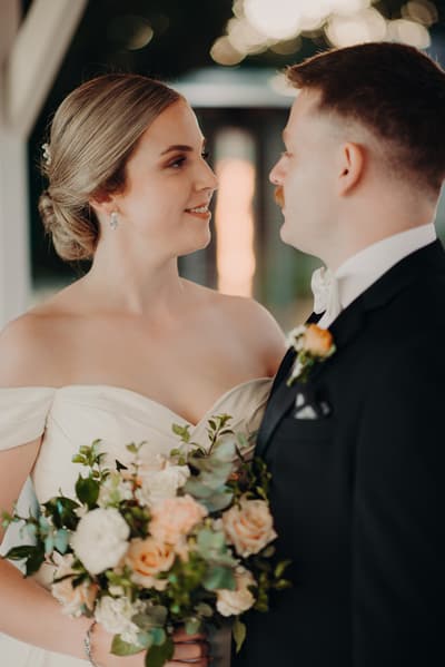 Courtney the bride in an off-shoulder white wedding gown holding a bouquet of flowers looks at Liam the groom in a black tuxedo with a white bow tie and boutonniere. Elopement photography packages Brisbane by She Said Yes.