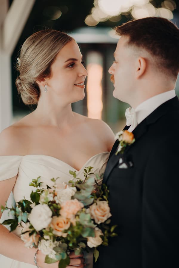 Courtney the bride in an off-shoulder white wedding gown holding a bouquet of flowers looks at Liam the groom in a black tuxedo with a white bow tie and boutonniere at Tiffany's Maleny.