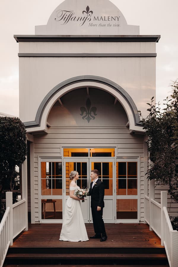 Courtney the bride and Liam the groom stand facing each other on the wooden steps outside Tiffany's Maleny, with the venue's entrance and signage visible behind them.