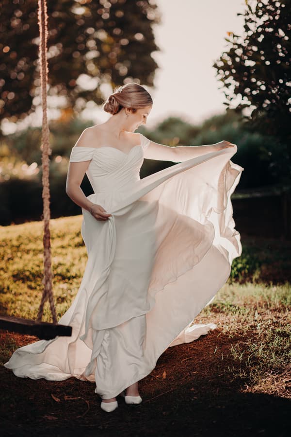 The bride Courtney poses alone outdoors at Tiffany's Maleny, holding and swirling the skirt of her off-shoulder wedding gown near a rope swing.
