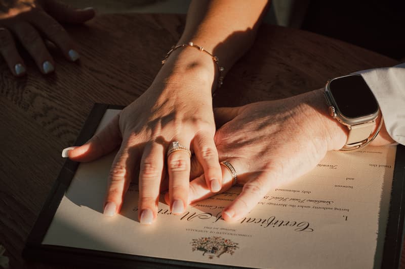 The hands of the brides, Mandy and Selina, rest on a wedding certificate at Sandstone Point Hotel — The Pavilion, showing their wedding rings.