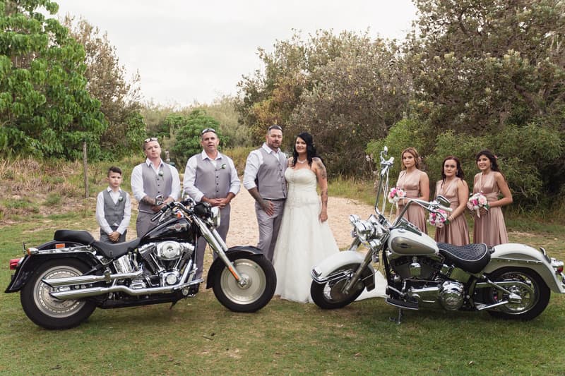 Kelly the bride and Shane the groom stand together outdoors at Novotel Sunshine Coast Resort with their bridal party, including three bridesmaids holding bouquets and three groomsmen in matching vests, posed between two motorcycles on grass with trees in the background.