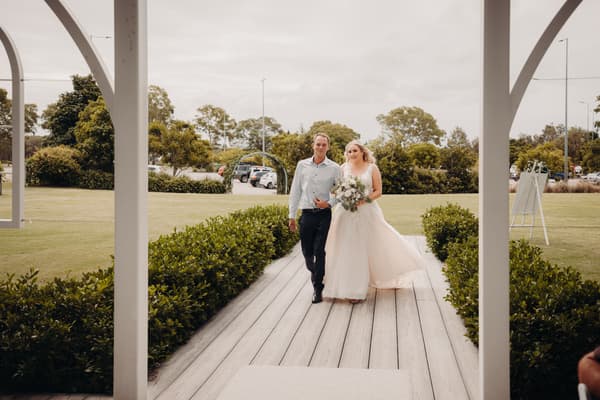 The bride Chloe and the groom Brodie walk together down a wooden aisle at Sandstone Point Hotel — Pavilion, surrounded by greenery and parked cars in the background.
