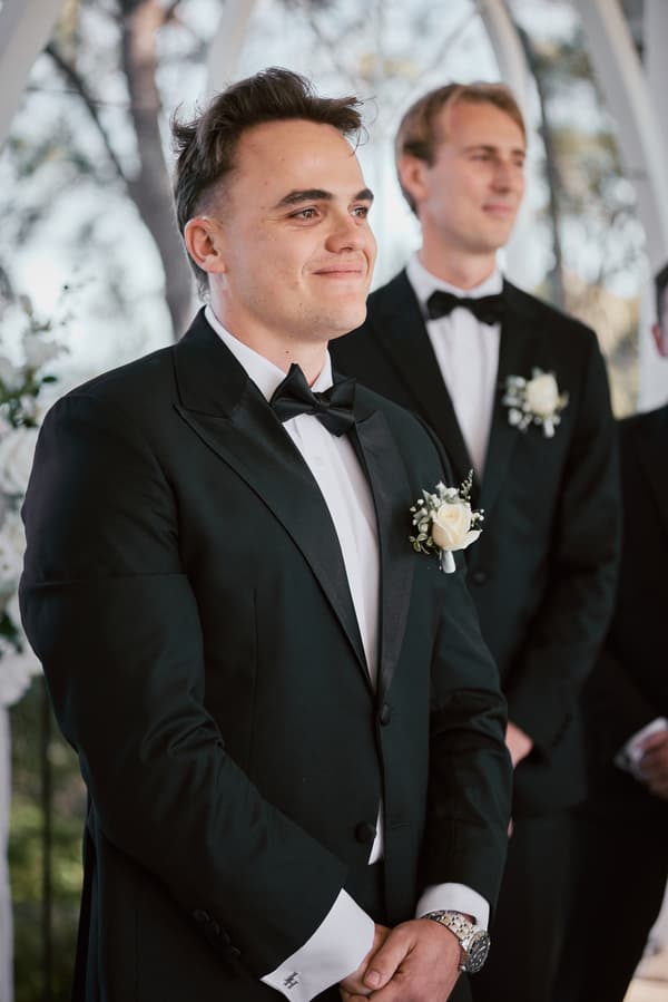 James, the groom, stands at the ceremony stage at Sandstone Point Hotel — The Pavilion, wearing a black tuxedo with a white rose boutonniere, accompanied by groomsmen also in black tuxedos with boutonnieres.