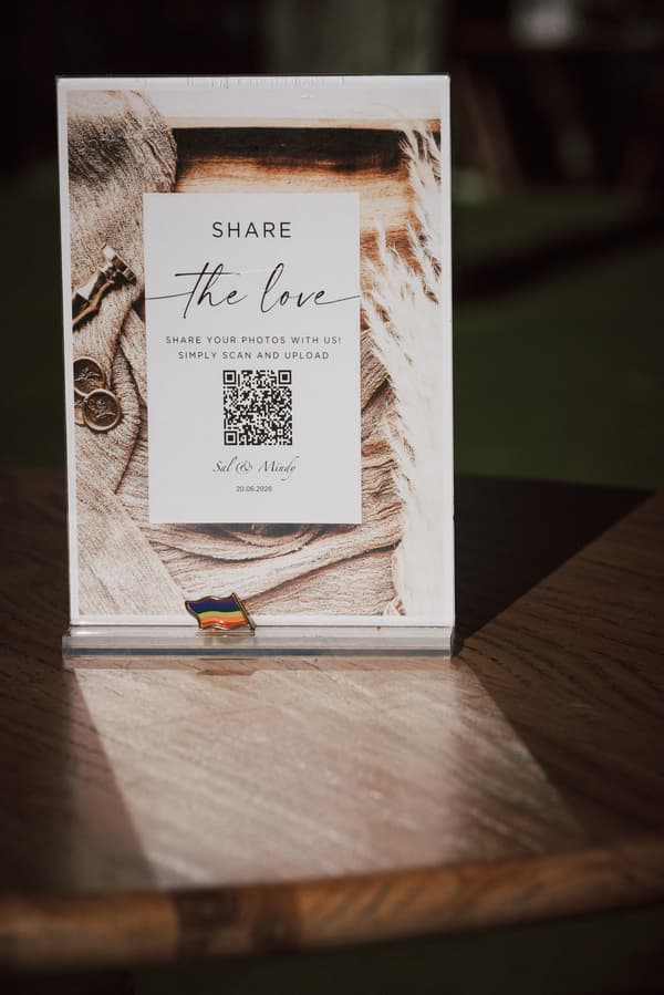 A table sign at Sandstone Point Hotel — The Pavilion invites guests to share photos by scanning a QR code, featuring the names Sal & Mandy and the date 20.06.2026, with a small rainbow flag pin at the base.