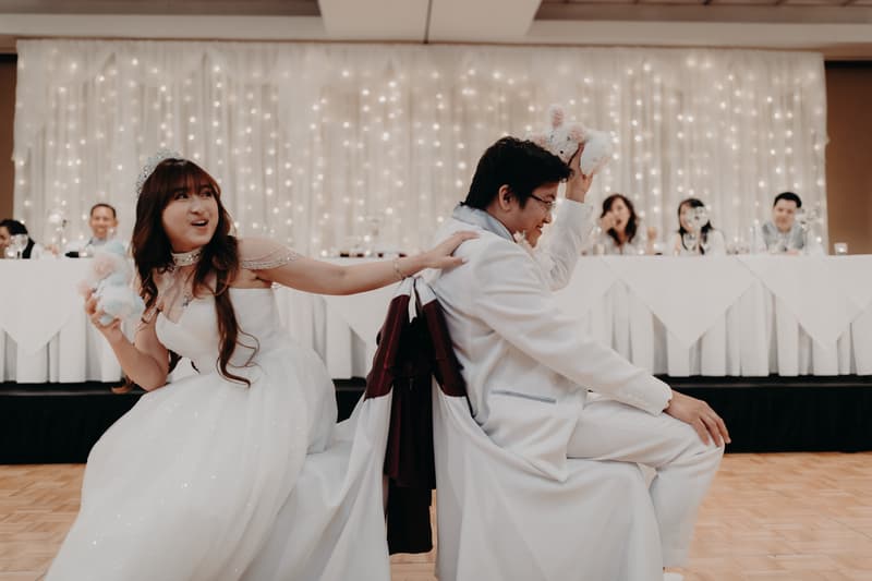 Bride Henny and groom Timothy sit back-to-back holding plush toys during a reception game at Royal on the Park, with the wedding party seated at a long table behind them against a backdrop of string lights.