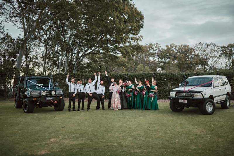 The bridal party poses outdoors on grass between two decorated 4WD vehicles at Sandstone Point Hotel — The Pavilion, with groomsmen in white shirts and black pants and bridesmaids in green dresses holding bouquets.