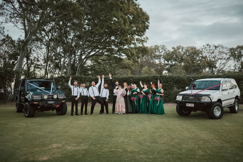 The bridal party poses outdoors on grass between two decorated 4WD vehicles, with groomsmen in white shirts and black pants and bridesmaids in green dresses holding bouquets. Wedding photographers in Brisbane by She Said Yes.