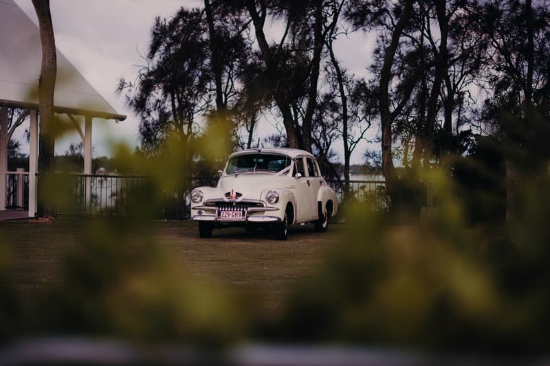 A vintage white car decorated with wedding ribbons is parked on the lawn near the Sandstone Point Hotel pavilion, surrounded by trees and greenery.
