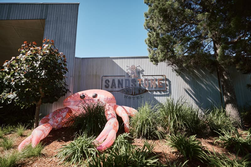 Exterior view of Sandstone Point Hotel with a large pink crab sculpture surrounded by plants and trees under a clear blue sky.