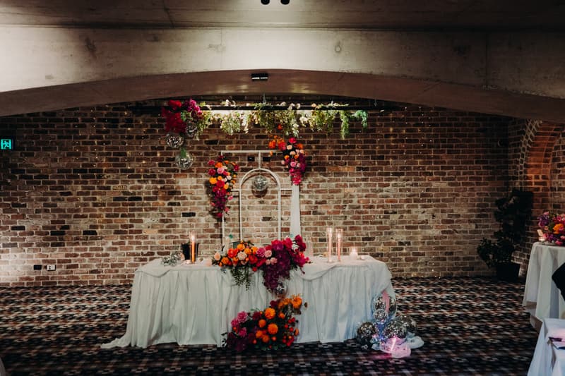 Reception stage at Sandstone Point Hotel — Cellar with a decorated table draped in white cloth, adorned with vibrant floral arrangements and candles against a brick wall backdrop.